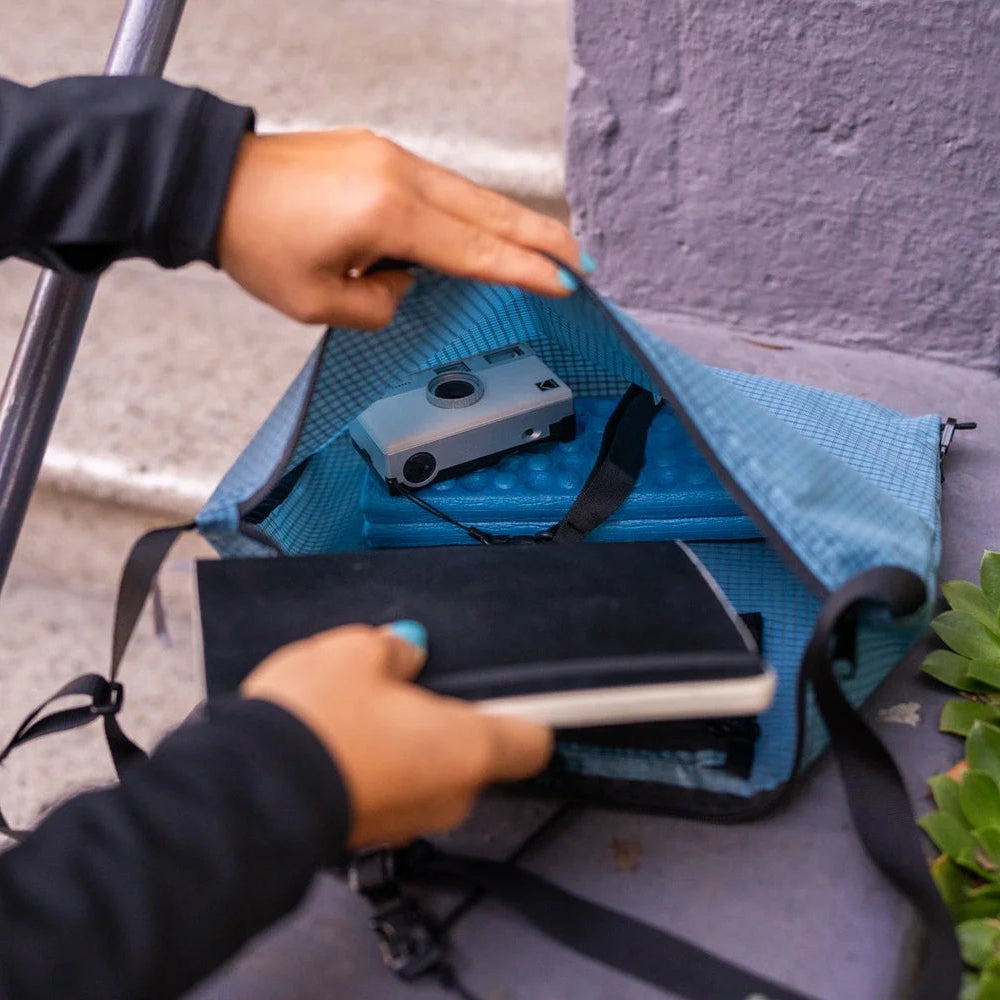 Person opening a blue smoke magic musette bag on a concrete surface with a camera and notebook inside
