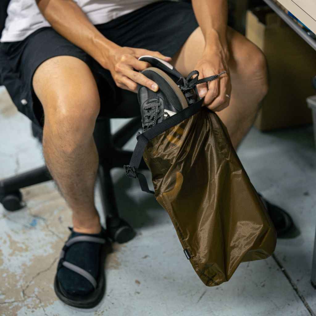 Person sitting on a chair stuffing a pair of shoes into a brown Fairweather Dry Sack .